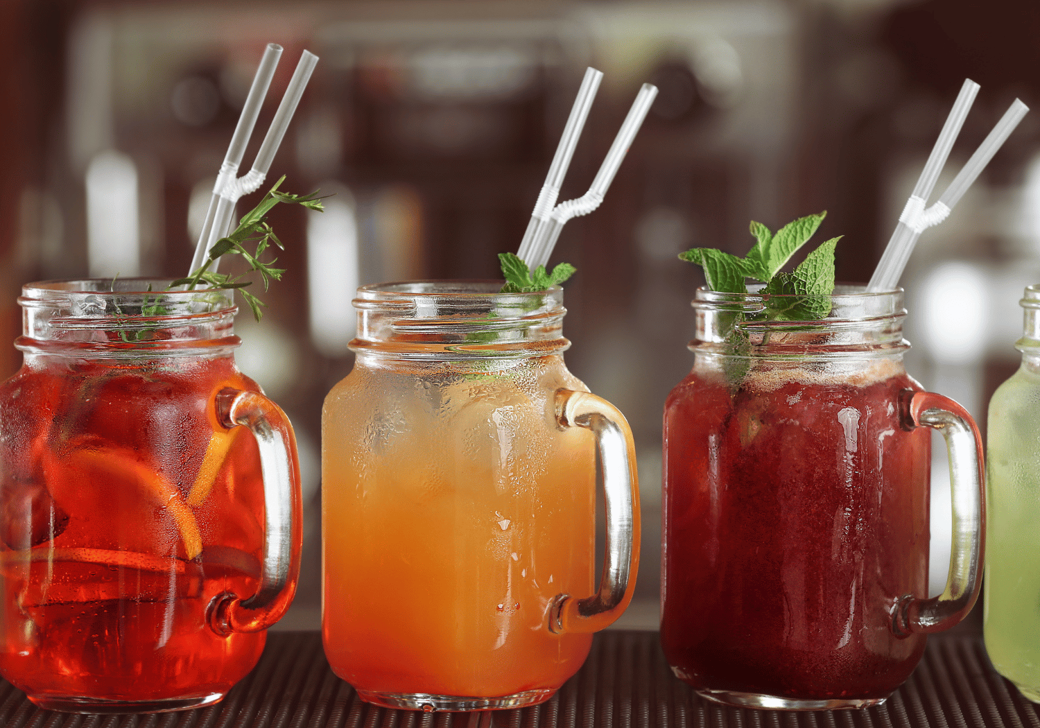Three mason jar drinks with mint garnish and white striped straws on a bar.